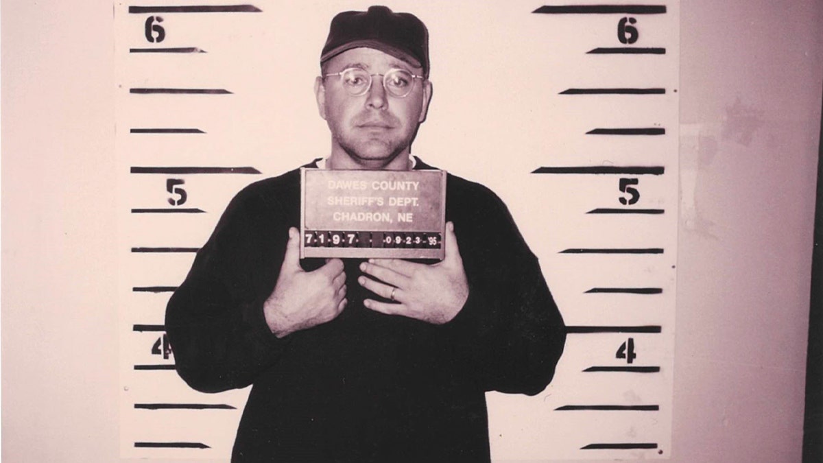 Tim Walz holding up a plate and standing in front of a wall with height markers in a 1995 mugshot